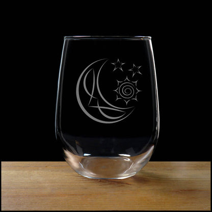 A clear stemless wine glass with an etched crescent moon and stars design, resting on a wooden surface against a solid black background.