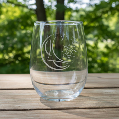 A clear stemless wine glass with an etched crescent moon and stars design, resting on a wooden surface against trees in the background.