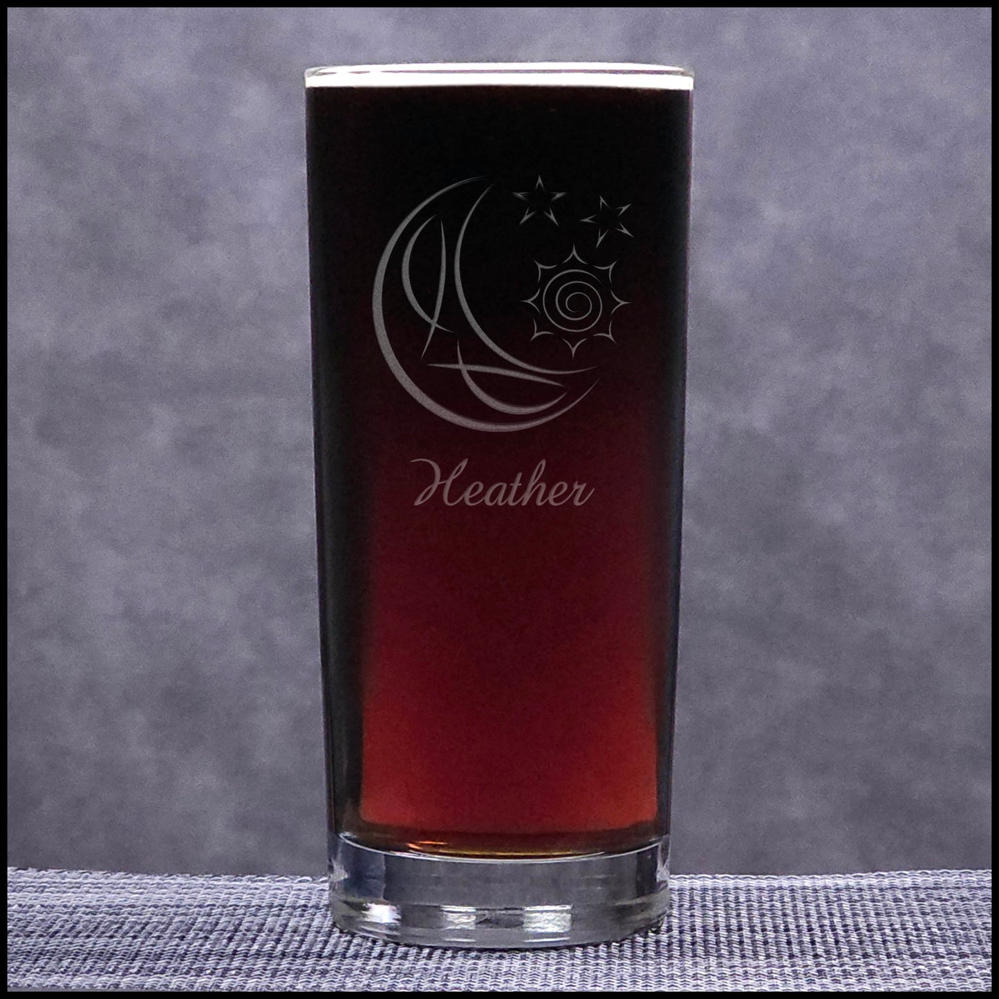 A clear cooler glass filled with a dark red-brown liquid, featuring a frosted crescent moon and sun design with the name Heather, set against a textured gray background.