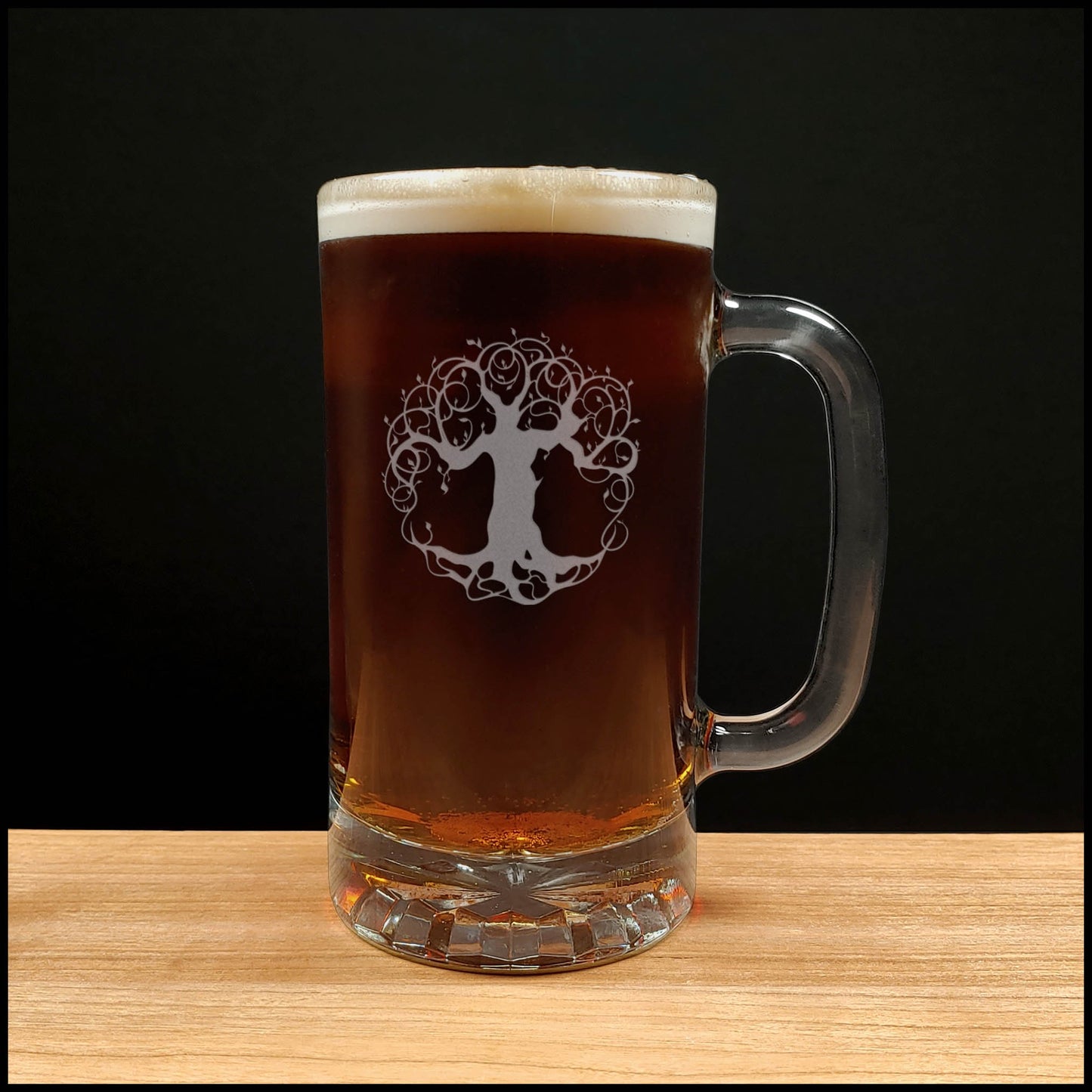 A clear glass beer mug with a handle, filled with dark amber beer and topped with white foam, featuring an etched tree-of-life graphic on the front, set on a light wood surface against a black background.