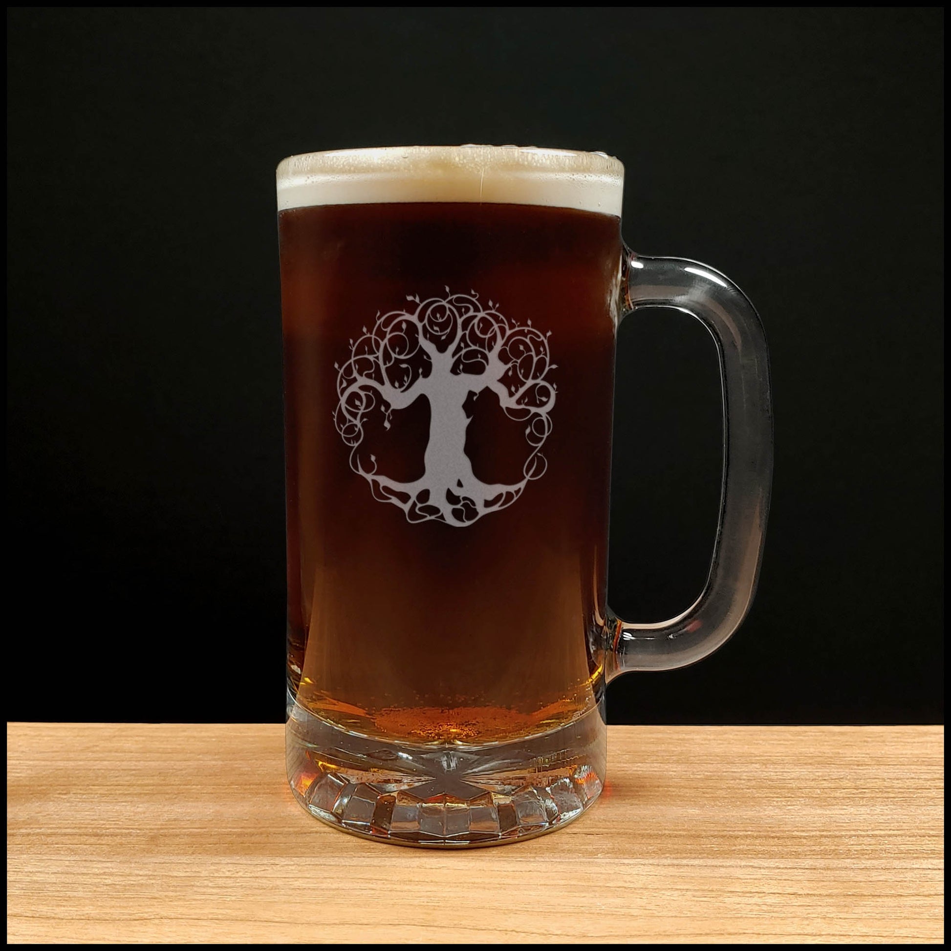 A clear glass beer mug with a handle, filled with dark amber beer and topped with white foam, featuring an etched tree-of-life graphic on the front, set on a light wood surface against a black background.