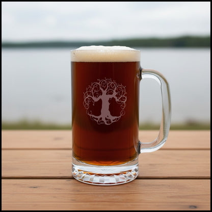 A clear glass beer mug filled with amber beer and topped with white foam, standing on a wooden table outdoors with a blurred lake and trees in the background. The mug has a frosted tree-of-life style graphic on the front.