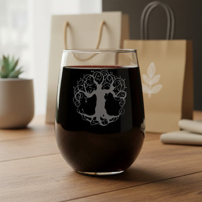 A stemless wine glass filled with red wine, featuring an etched tree-of-life design on the front, set on a wooden table with gift bags in the background.