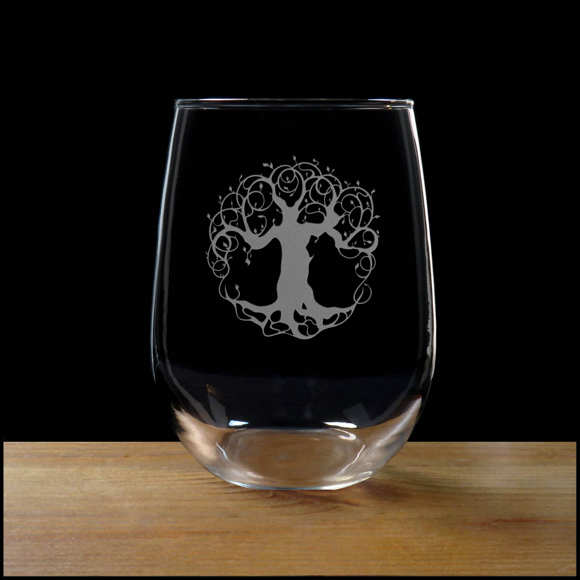 A clear stemless wine glass with a silver etched tree of life design, resting on a wooden surface against a solid black background.