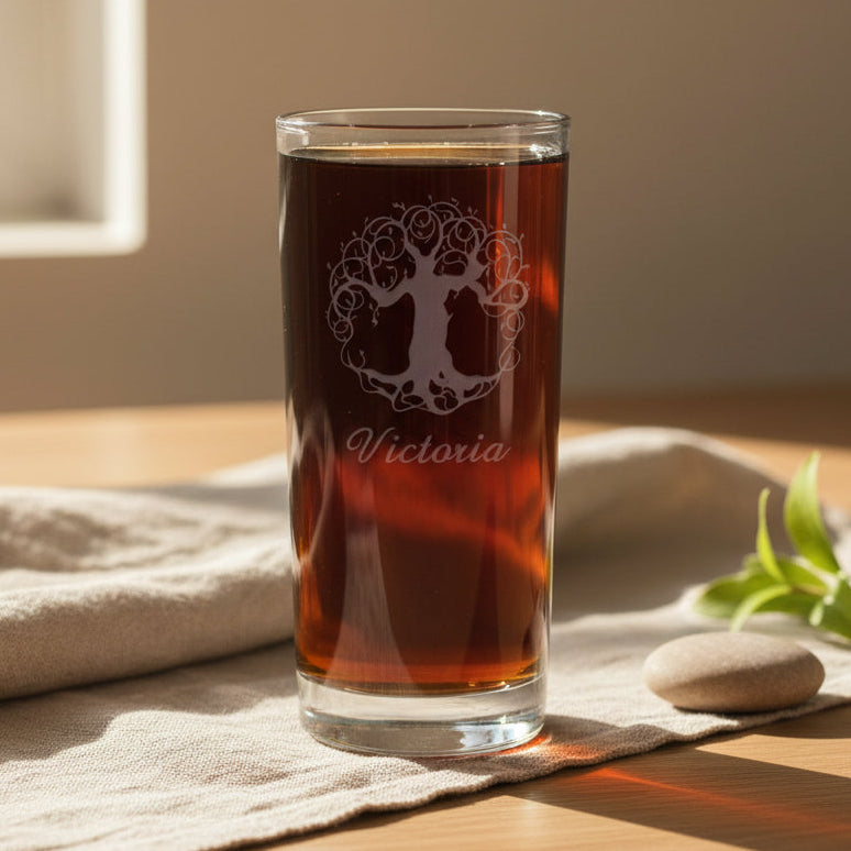 A clear glass tumbler filled with amber-brown liquid, standing on a beige cloth on a wooden surface. The glass has a white tree-of-life style graphic and the name 'Victoria' printed on it.