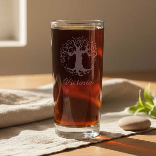 A clear glass tumbler filled with amber-brown liquid, standing on a beige cloth on a wooden surface. The glass has a white tree-of-life style graphic and the name 'Victoria' printed on it.