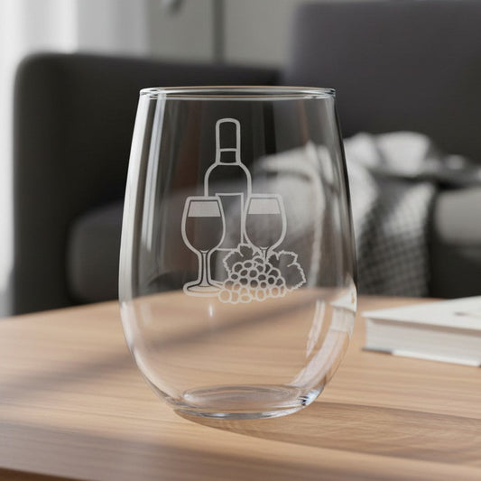 A clear stemless wine glass with an etched wine bottle, two wine glasses, and a grape cluster design, sitting on a light wood table in a blurred living room setting.
