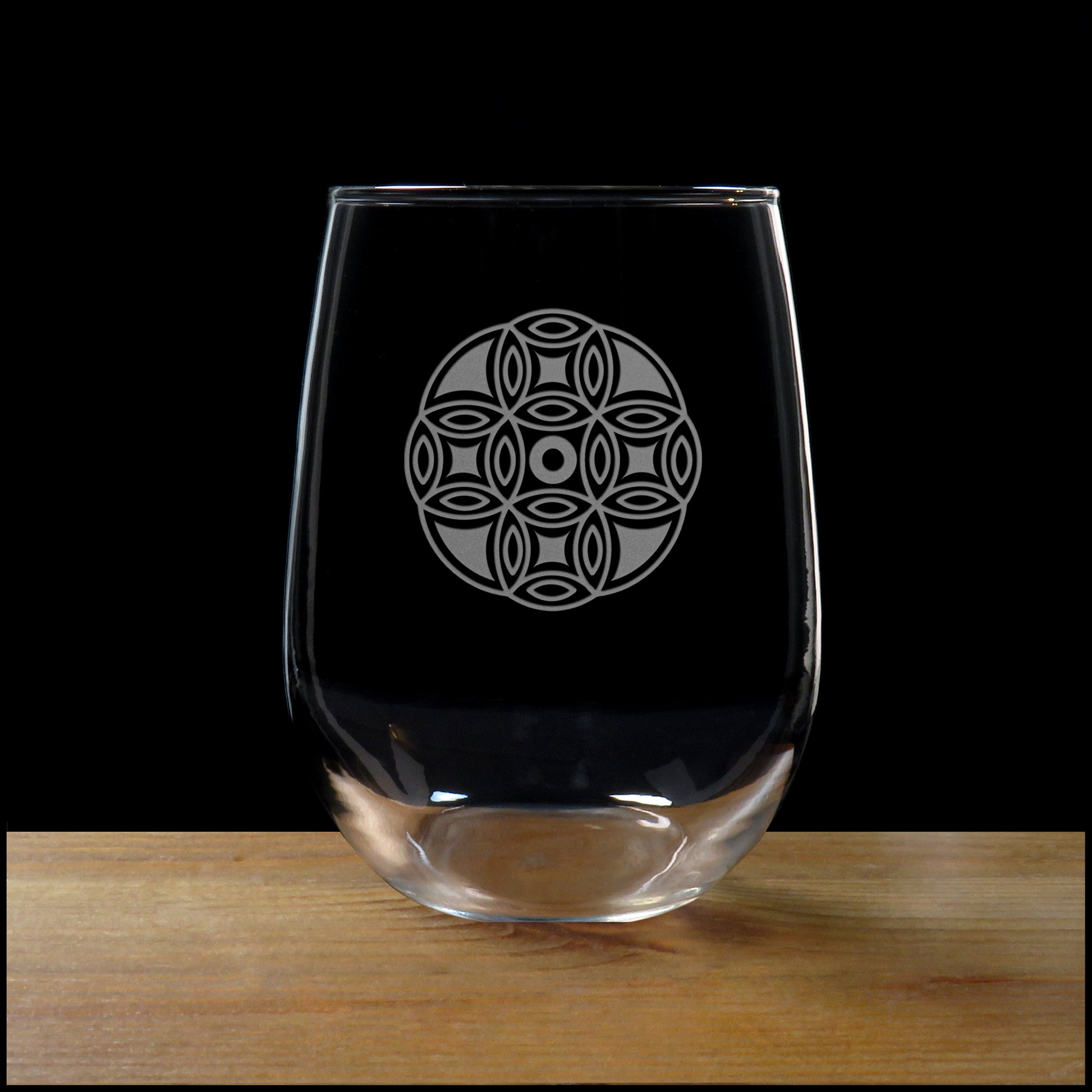 A clear stemless wine glass with a etched mandala etched design on the front, shown standing on a wooden surface against a black background.