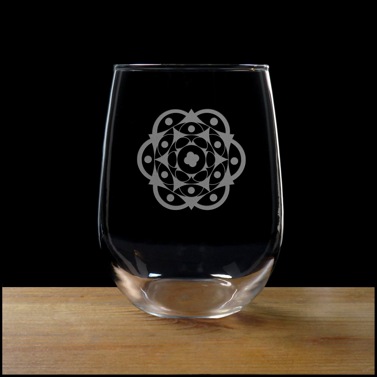 A clear stemless wine glass with a etched mandala design printed on the front, resting on a wooden surface against a solid black background.