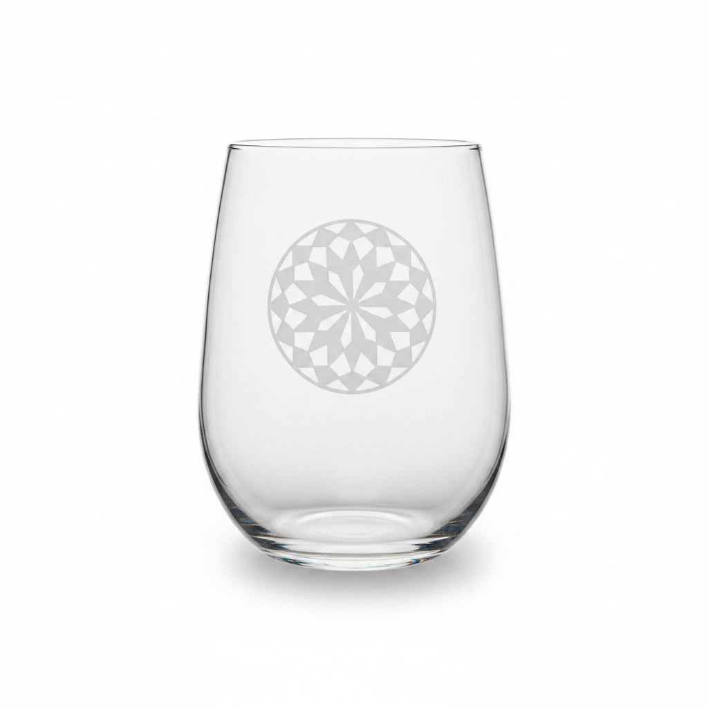 A clear stemless wine glass with a mandala design sand etched on the front, resting on a wooden surface against a white background.