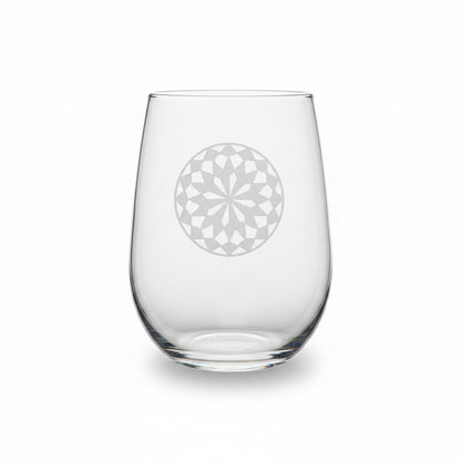 A clear stemless wine glass with a mandala design sand etched on the front, resting on a wooden surface against a white background.