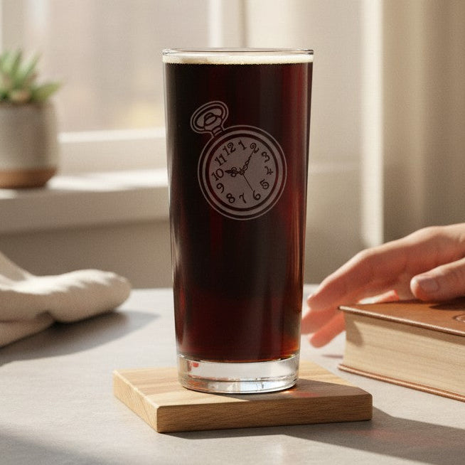 A clear highball glass filled with dark brown beer, with an etched poket watch design sits on a small wooden coaster on a table. A person's hands are resting on a brown book nearby, with a window, curtains, and a small potted plant in the background.