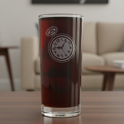 A tall clear drinking glass filled with a dark reddish-brown liquid, standing on a wooden table in a softly blurred living room. The glass has an etched pocket-watch design on the front.
