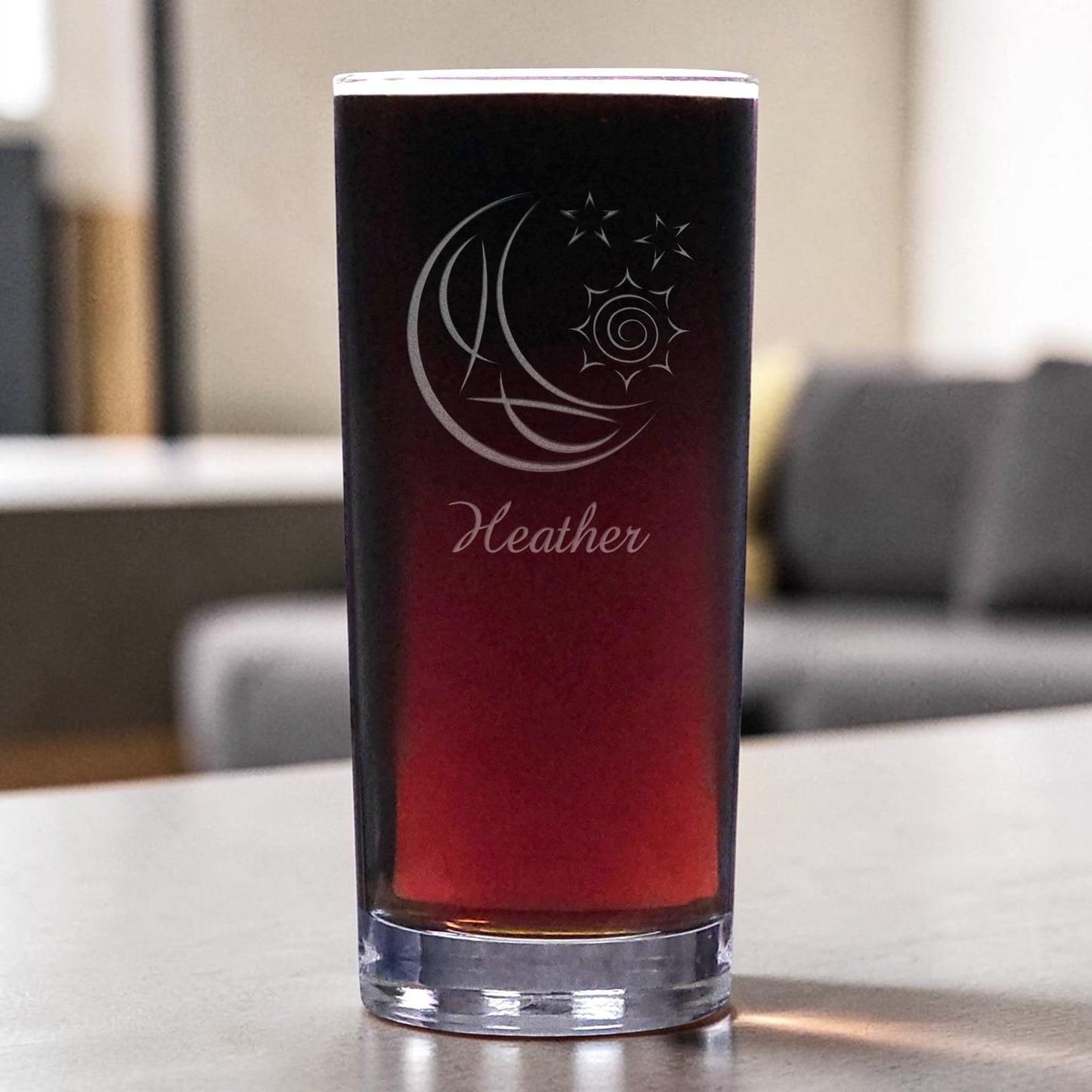 A clear pint glass filled with a dark red drink, featuring an etched crescent moon and sun design with the name 'Heather' below it, set on a tabletop with a blurred living room background.