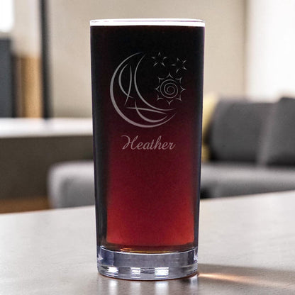 A clear pint glass filled with a dark red drink, featuring an etched crescent moon and sun design with the name 'Heather' below it, set on a tabletop with a blurred living room background.