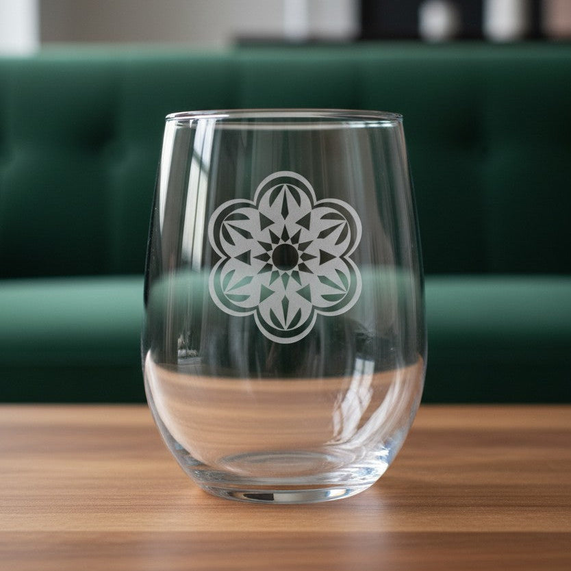 A clear stemless wine glass with a white geometric floral pattern on the front, sitting on a wooden table with a blurred green sofa in the background.