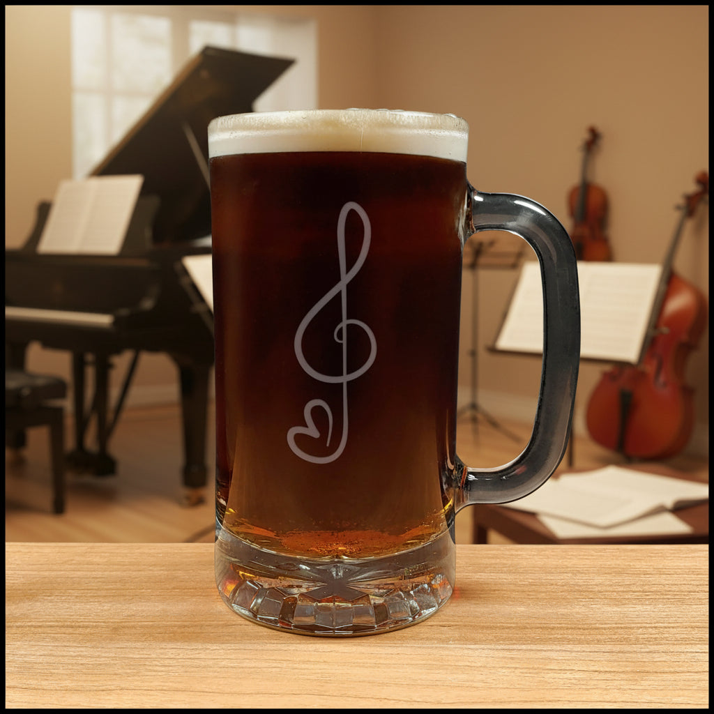 A clear glass beer mug with a handle, filled with amber beer and topped with white foam, featuring a etched treble clef and heart engraving on the front. The mug sits on a wooden table with a blurred dark green couch in the background.