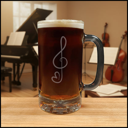 A clear glass beer mug with a handle, filled with amber beer and topped with white foam, featuring a etched treble clef and heart engraving on the front. The mug sits on a wooden table with a blurred dark green couch in the background.