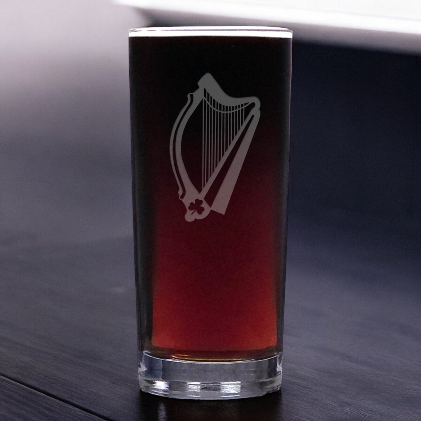 A clear glass pint glass with an etched harp design, filled with a dark red beverage, set on a dark wooden surface.