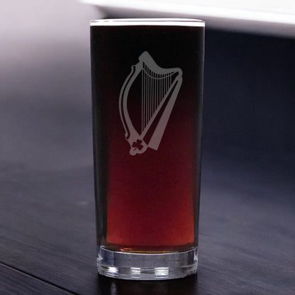 A clear glass pint glass with an etched harp design, filled with a dark red beverage, set on a dark wooden surface.