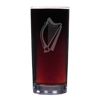 A clear pint glass filled with dark red beer, featuring a white etched harp design on the front, set against a plain white background.
