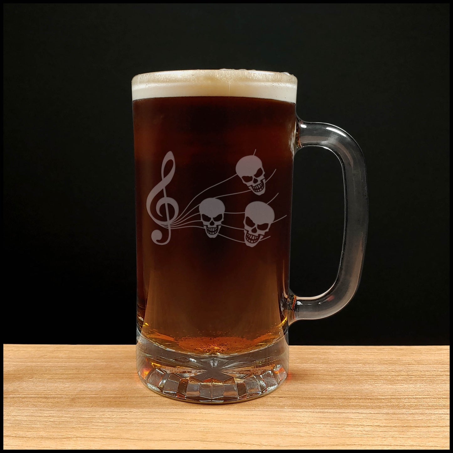 A clear glass beer mug with a handle, filled with dark red beer and topped with white foam, featuring a white etched design of a treble clef and three skulls. The mug is placed on a light wood surface against a solid black background.