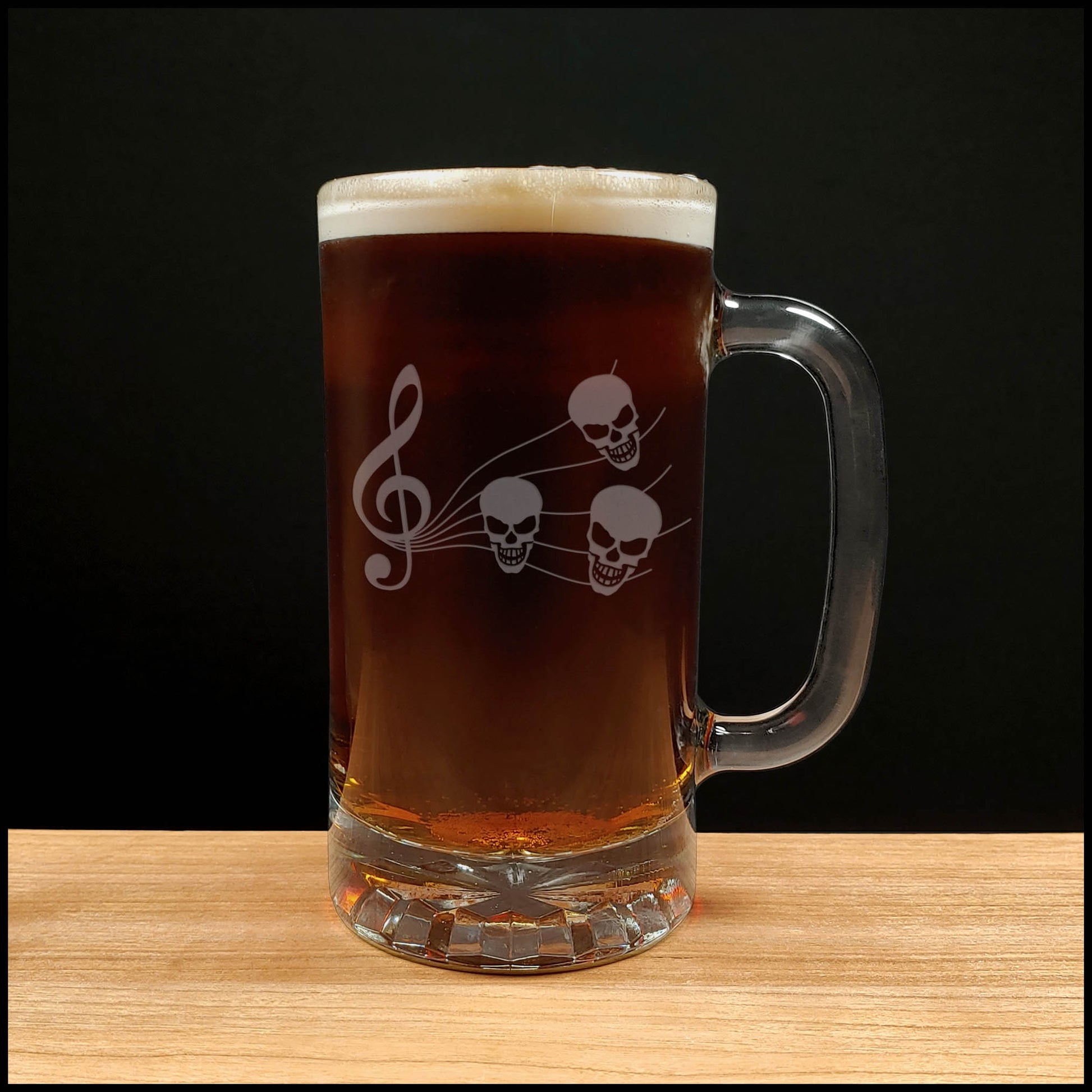 A clear glass beer mug with a handle, filled with dark red beer and topped with white foam, featuring a white etched design of a treble clef and three skulls. The mug is placed on a light wood surface against a solid black background.