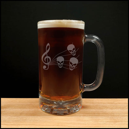 A clear glass beer mug with a handle, filled with dark red beer and topped with white foam, featuring a white etched design of a treble clef and three skulls. The mug is placed on a light wood surface against a solid black background.