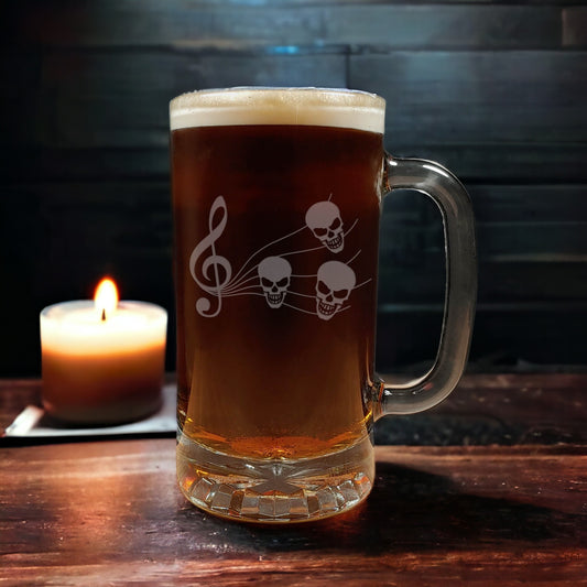 A clear glass beer mug with a handle, filled with amber beer and topped with white foam, featuring a frosted music staff design of a treble clef and three skulls. The mug sits on a dark wooden table next to a lit candle against a dark wood-paneled background.