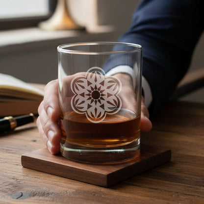 A hand holds a clear rocks glass with a white floral etched mandala design, containing amber whiskey, resting on a square wooden coaster at a wooden table. A person in a navy suit sleeve is visible on the right, with an open book and pen in the background.