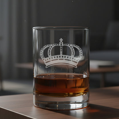 A clear rocks glass with a white crown engraving, partially filled with amber-colored liquor, set on a light wood surface 