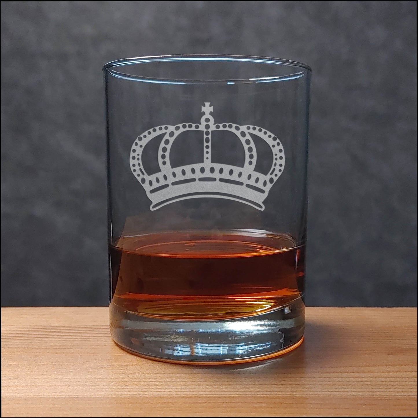 A clear rocks glass with a white crown engraving, partially filled with amber-colored liquor, set on a light wood surface against a dark gray textured background.