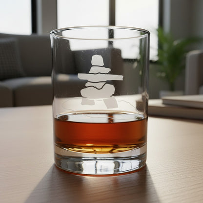 A clear rocks glass with a etched Inuksuk design, partially filled with amber whiskey, sitting on a light wood table