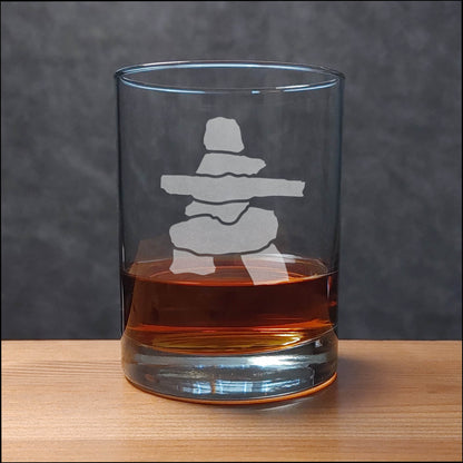 A clear glass rocks tumbler with an etched Inuksuk design, partially filled with amber whiskey, resting on a light wood surface against a dark gray textured background.