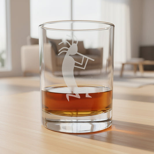 A clear rocks glass with an etched Kokopelli dancer design, partially filled with amber whiskey, sitting on a light wood surface against a living room