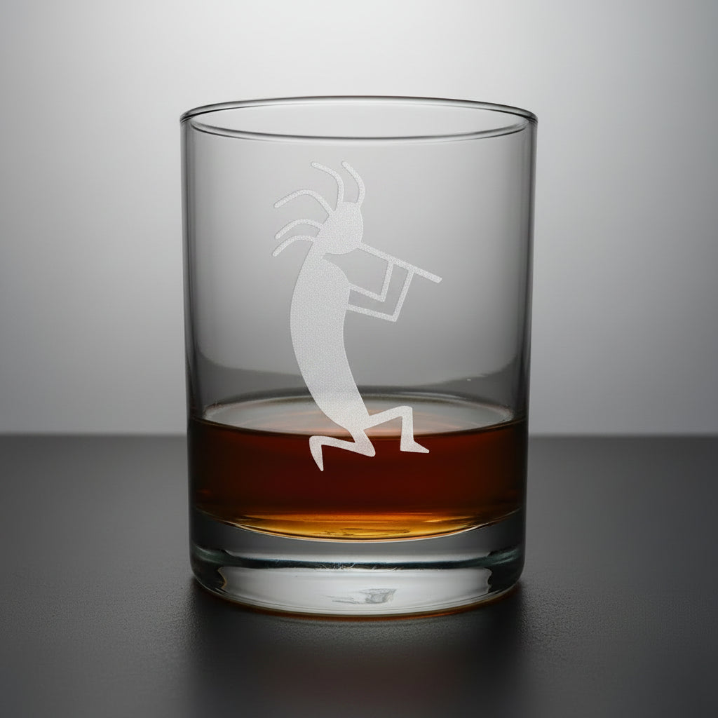 A clear rocks glass with an etched Kokopelli dancer design, partially filled with amber whiskey, sitting on a light wood surface against a gray background.