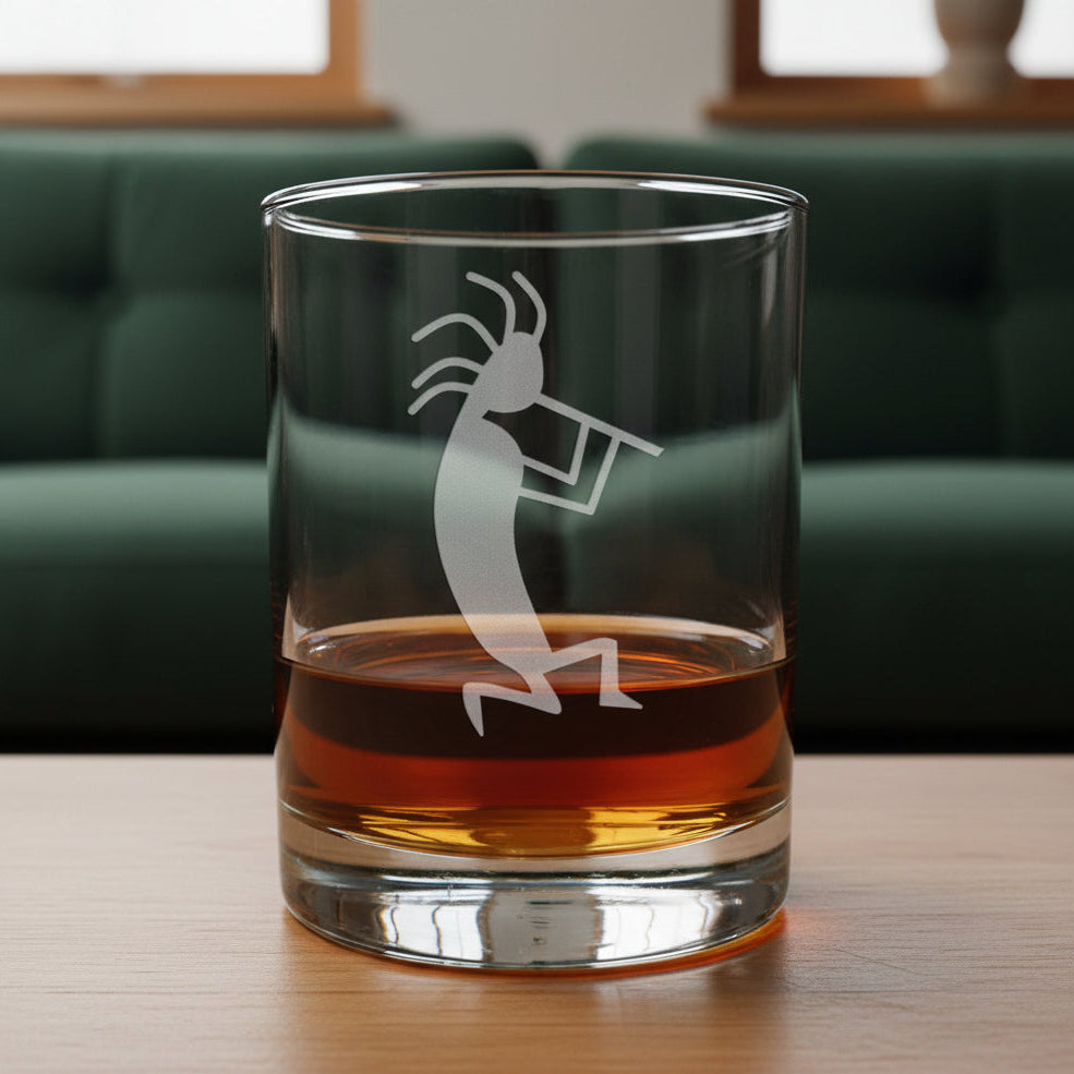 A clear rocks glass with an etched Kokopelli dancer design, partially filled with amber whiskey, sitting on a light wood surface against a dark green couch