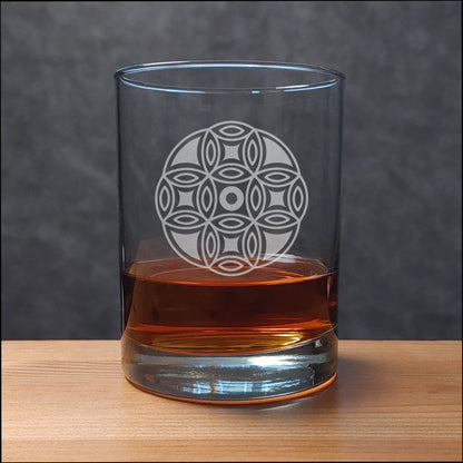 A clear glass rocks tumbler with an etched geometric flower pattern, partially filled with amber whiskey, set on a wooden surface against a dark gray background.