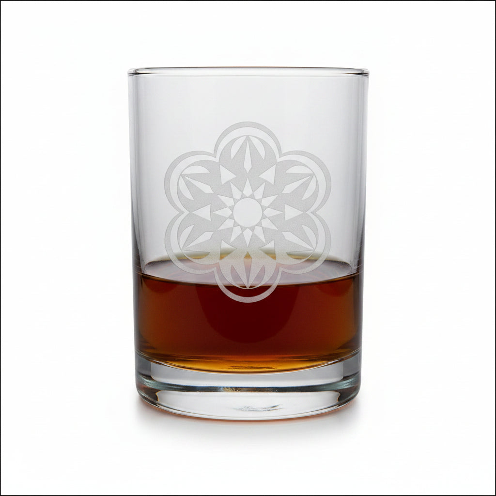 A clear rocks glass with an etched floral mandala design, partially filled with amber whiskey, set on a light wood surface against a white background.