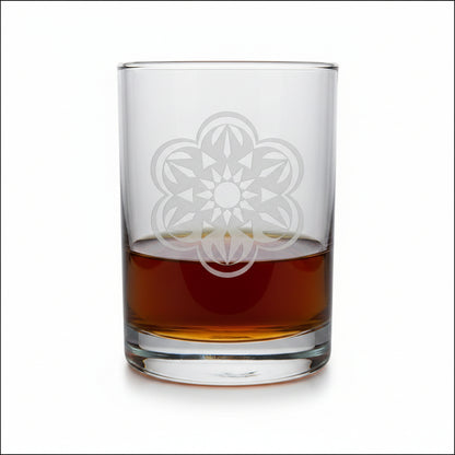 A clear rocks glass with an etched floral mandala design, partially filled with amber whiskey, set on a light wood surface against a white background.