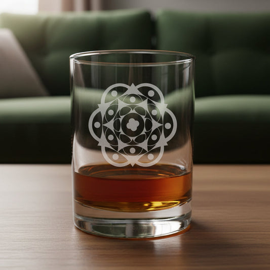 A clear glass rocks tumbler with an etched geometric mandala design, partially filled with amber-colored liquor, sitting on a light wood surface against a dark gray textured background.