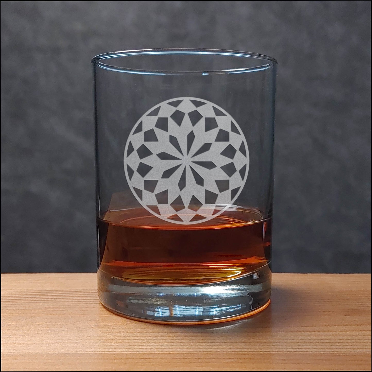 A clear rocks glass with an etched floral mandala design, partially filled with amber whiskey, set on a light wood surface against a dark gray textured background.