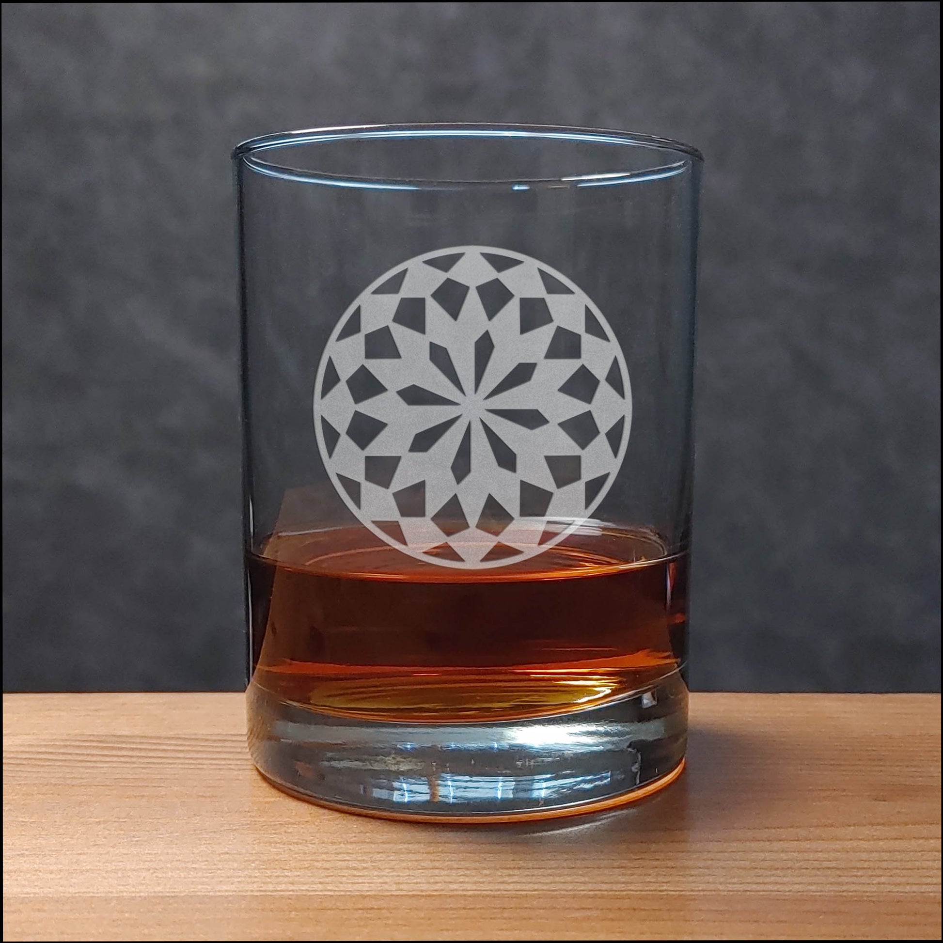 A clear rocks glass with an etched floral mandala design, partially filled with amber whiskey, set on a light wood surface against a dark gray textured background.