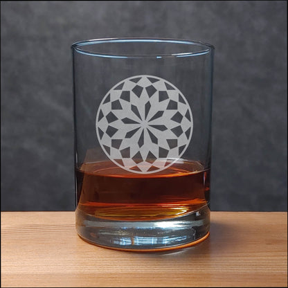 A clear rocks glass with an etched floral mandala design, partially filled with amber whiskey, set on a light wood surface against a dark gray textured background.