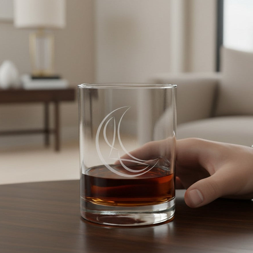 A clear lowball glass with an etched crescent moon design, partially filled with amber liquid, resting on a dark wooden table with a hand holding the side. The background shows a softly blurred modern living room interior.