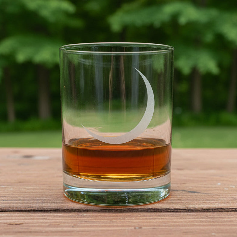 A clear glass rocks tumbler with an etched crescent moon design, partially filled with amber whiskey, set on a light wood surface against trees in the background.