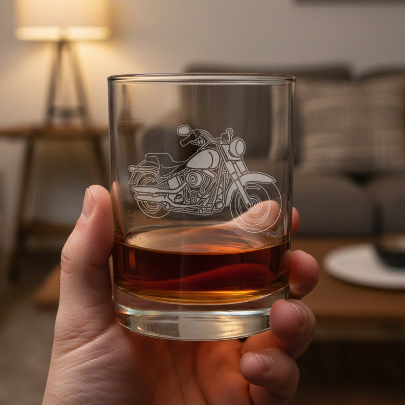 A hand holding a clear glass rocks tumbler with an etched motorcycle design, partially filled with an amber-colored drink