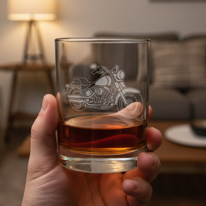 A hand holding a clear glass rocks tumbler with an etched motorcycle design, partially filled with an amber-colored drink
