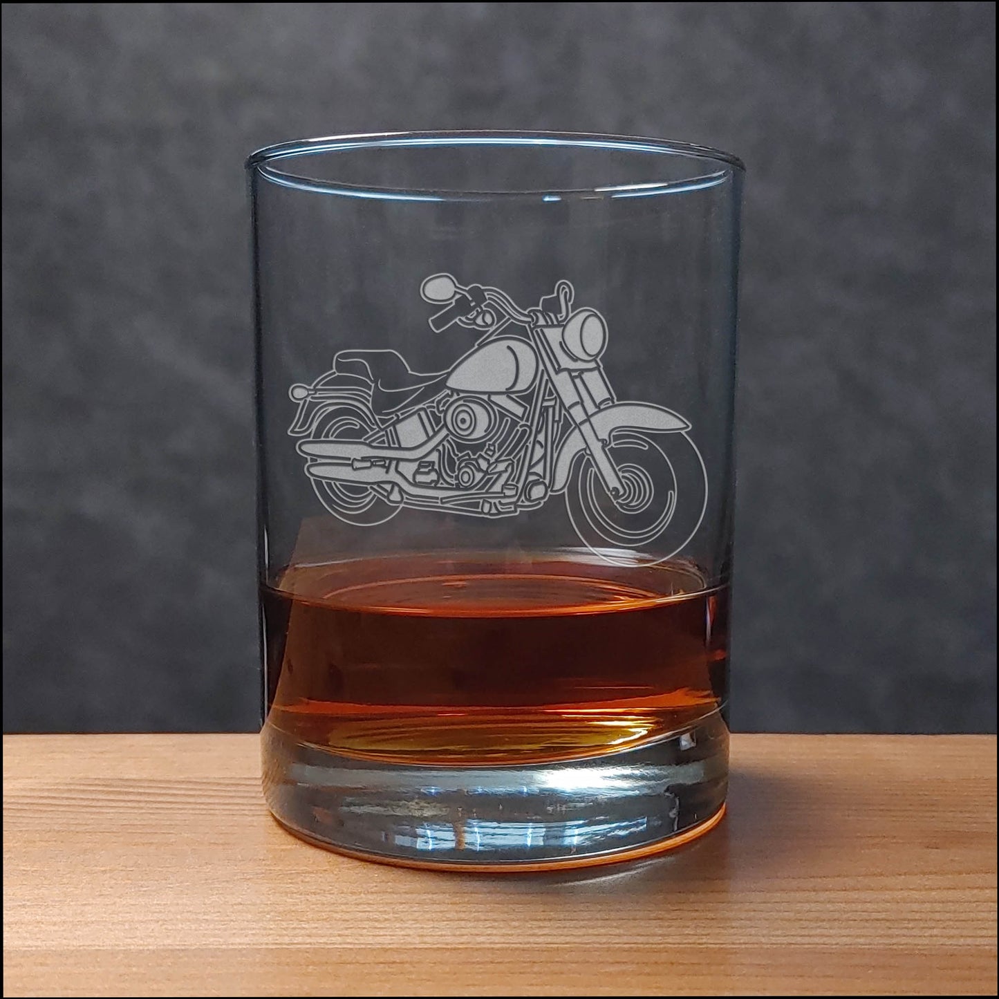 A clear glass rocks tumbler with an etched motorcycle design, partially filled with an amber-colored drink, set on a wooden surface against a dark gray textured background.