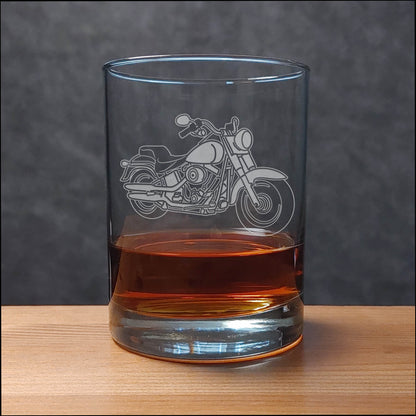 A clear glass rocks tumbler with an etched motorcycle design, partially filled with an amber-colored drink, set on a wooden surface against a dark gray textured background.